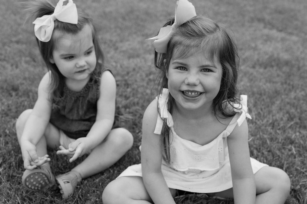 Two children sitting on grass in black and white, wearing bows in their hair and casual summer clothes.
