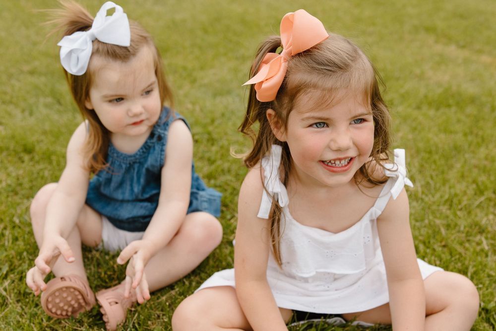 Two children sitting on grass wearing matching bows and summer outfits.