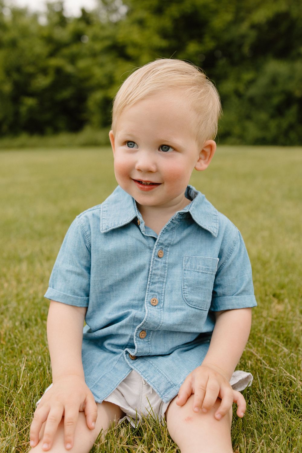 Toddler sitting on grass wearing blue button-up shirt smiles at camera during outdoor portrait session.