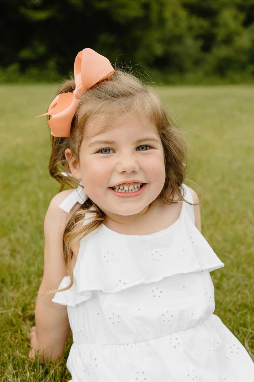 Child wearing white eyelet top and peach bow smiling in sunny field.