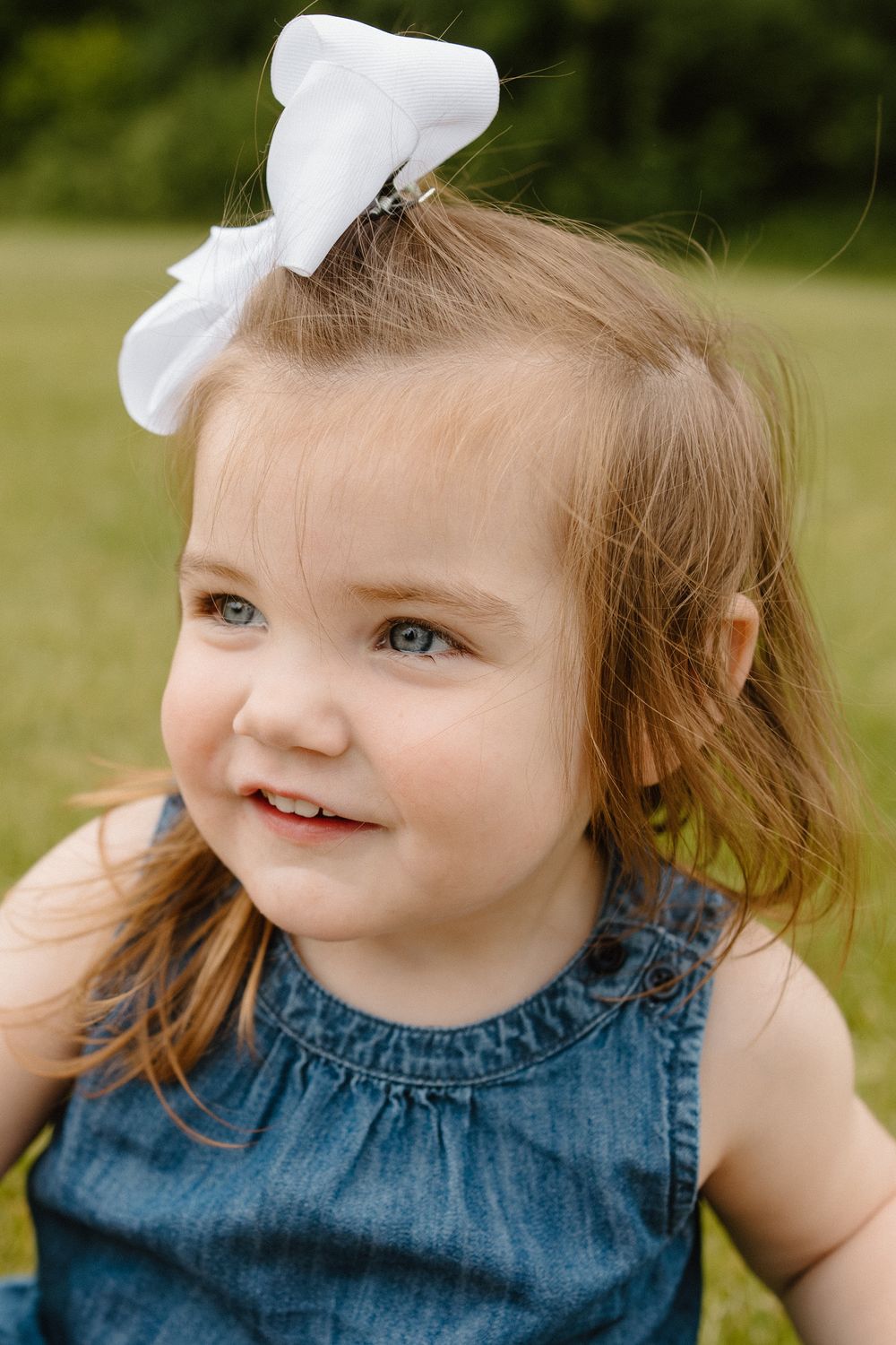 Young child in blue denim dress and white bow smiling outdoors.