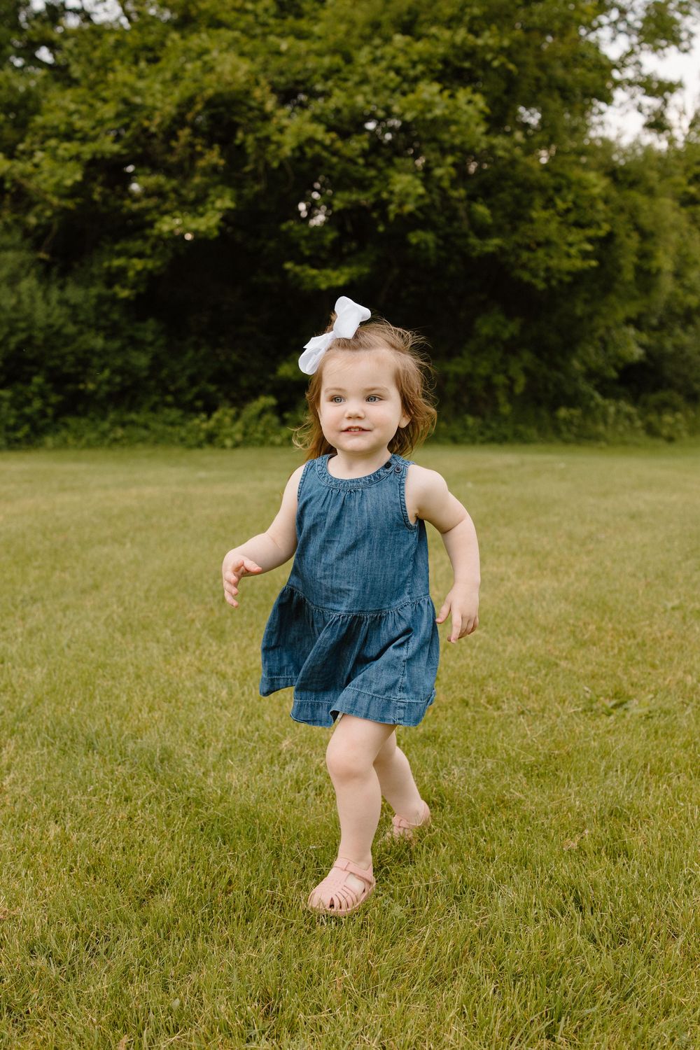 A toddler in a blue denim dress runs joyfully across a green lawn on a sunny day.