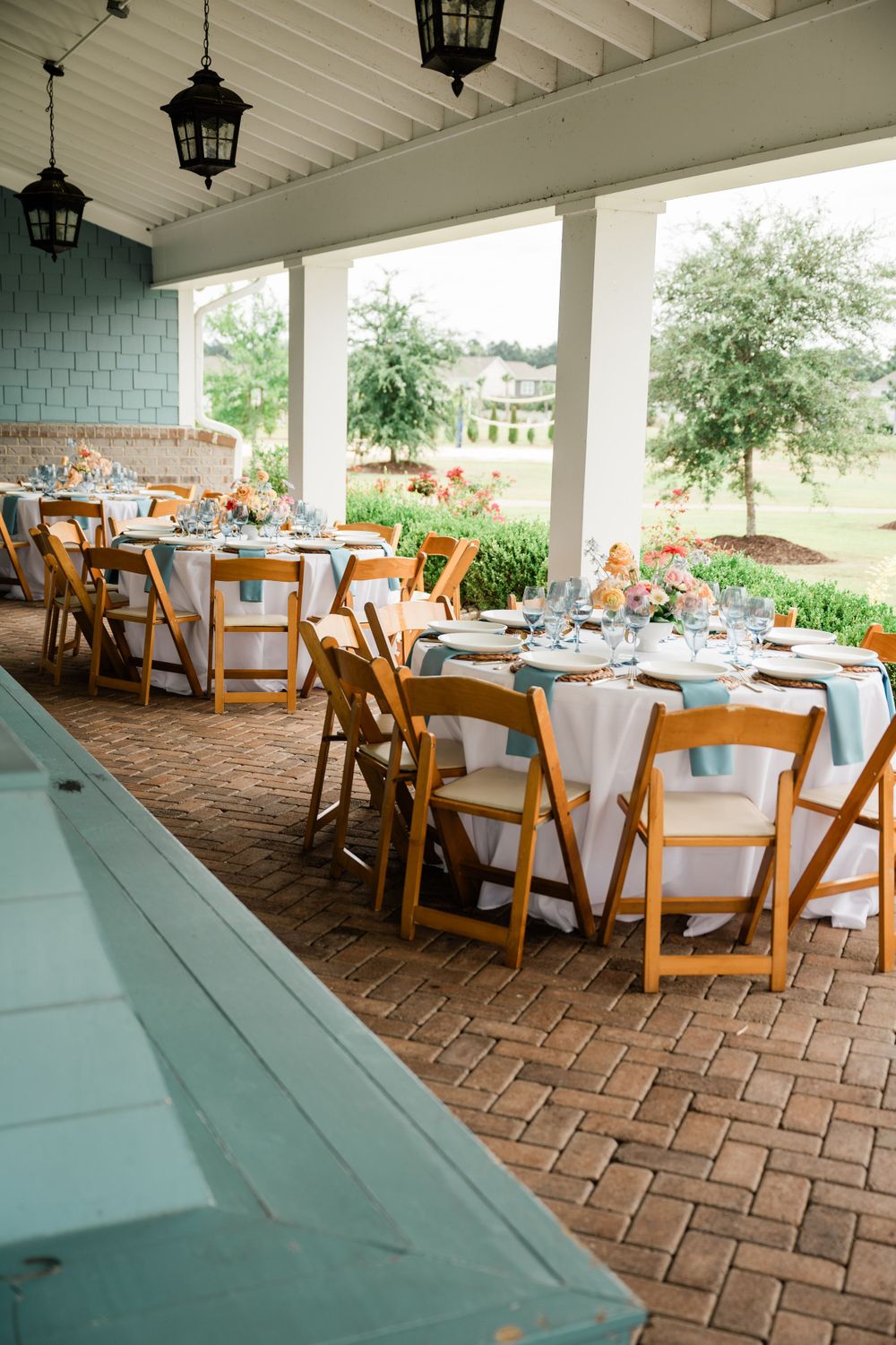 Elegant outdoor wedding reception setup with round tables and wooden chairs under a covered porch with hanging lanterns.