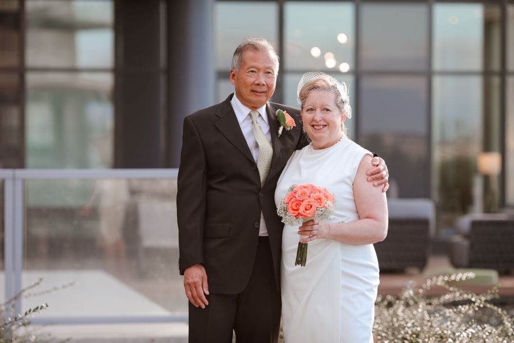 A bride in a white dress holds a coral bouquet while posing with her groom outdoors.