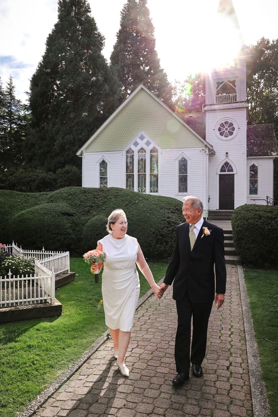 A wedding couple walks hand in hand on a brick path in front of a charming white chapel.
