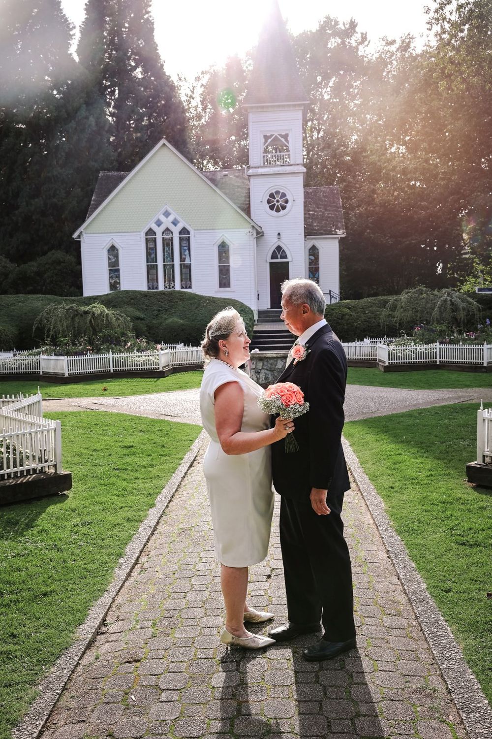 A bride and groom stand together on a brick pathway with a white chapel in the background.
