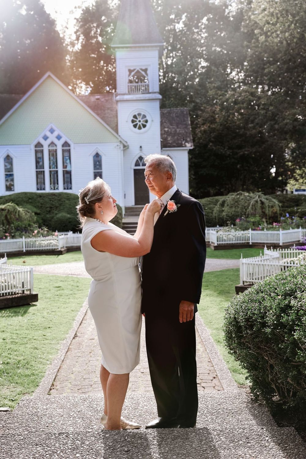A couple shares an intimate moment on a garden path with a historic white church behind them.