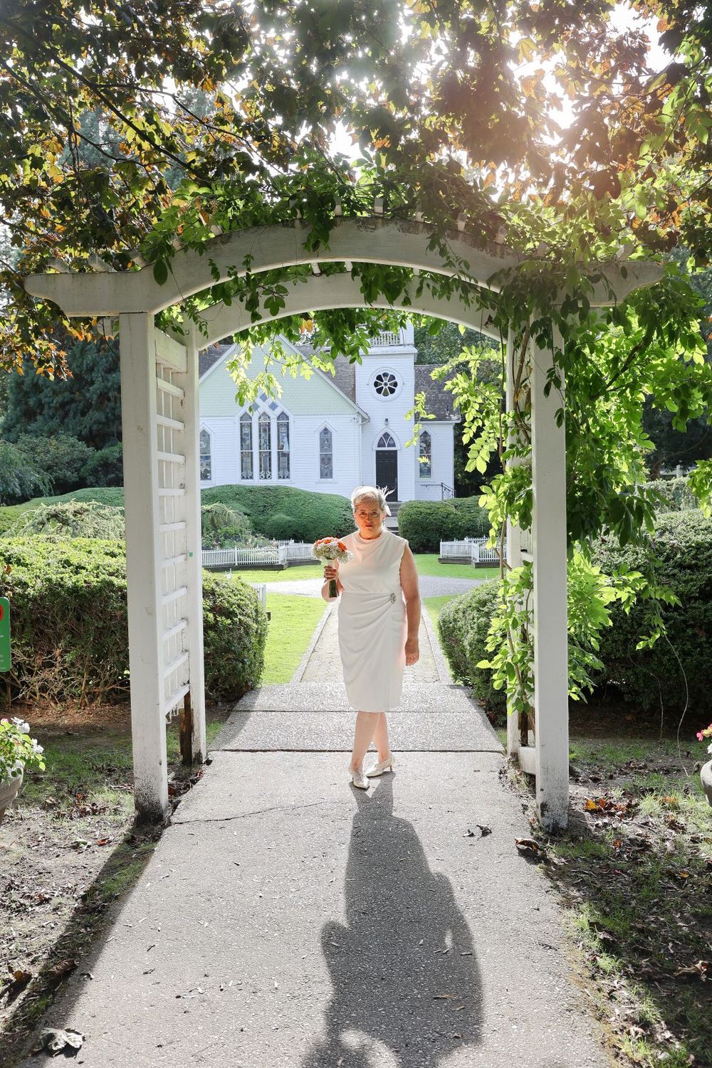 A person in white walks through a garden archway towards a quaint chapel.