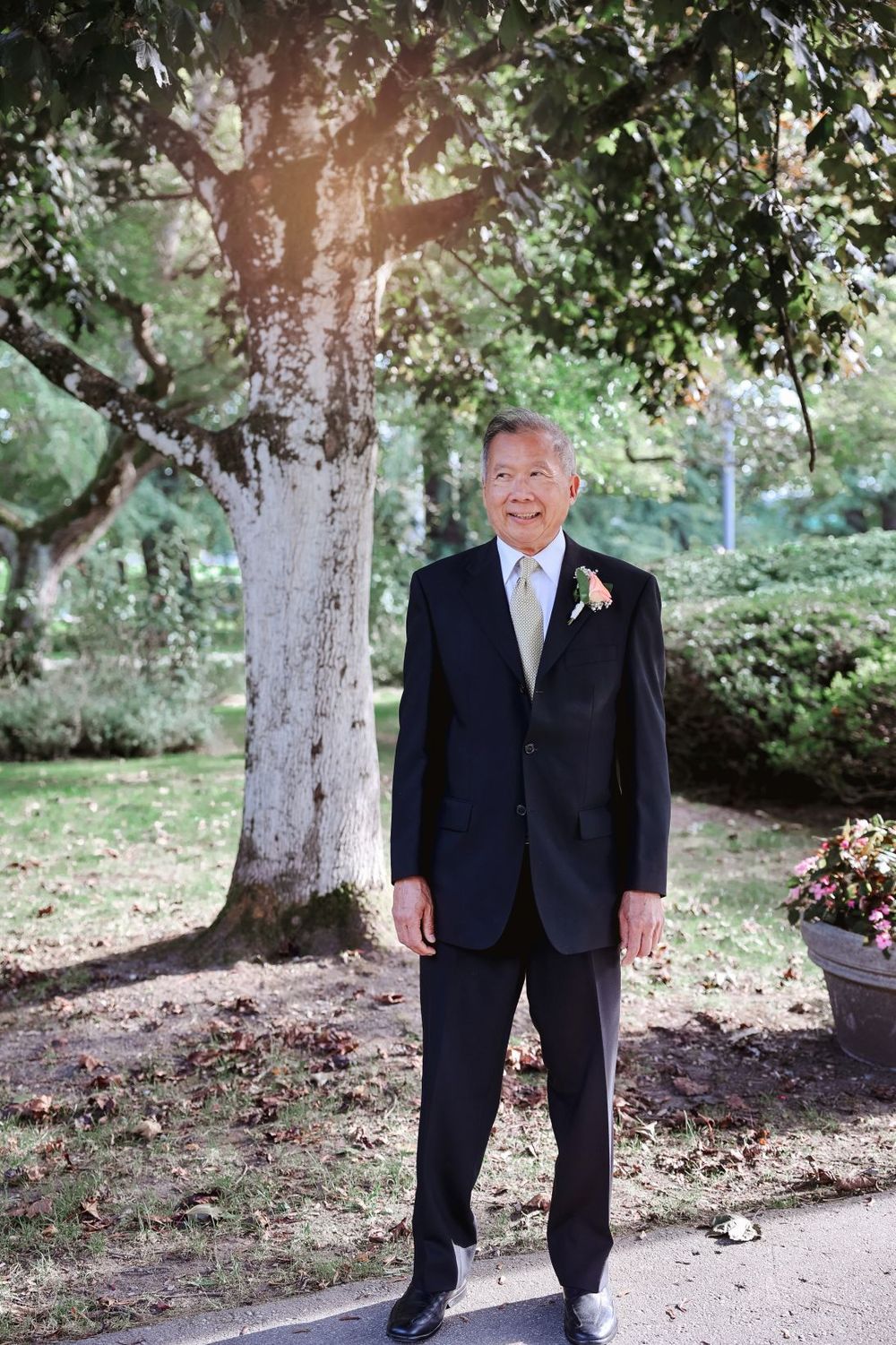 A groom in a navy suit poses outdoors under a sunlit tree.