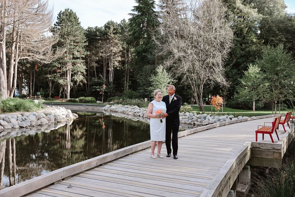 A couple stands together on a wooden bridge over a reflective pond.