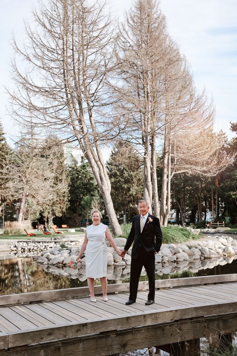 A couple holds hands while standing on a wooden bridge surrounded by bare winter trees.