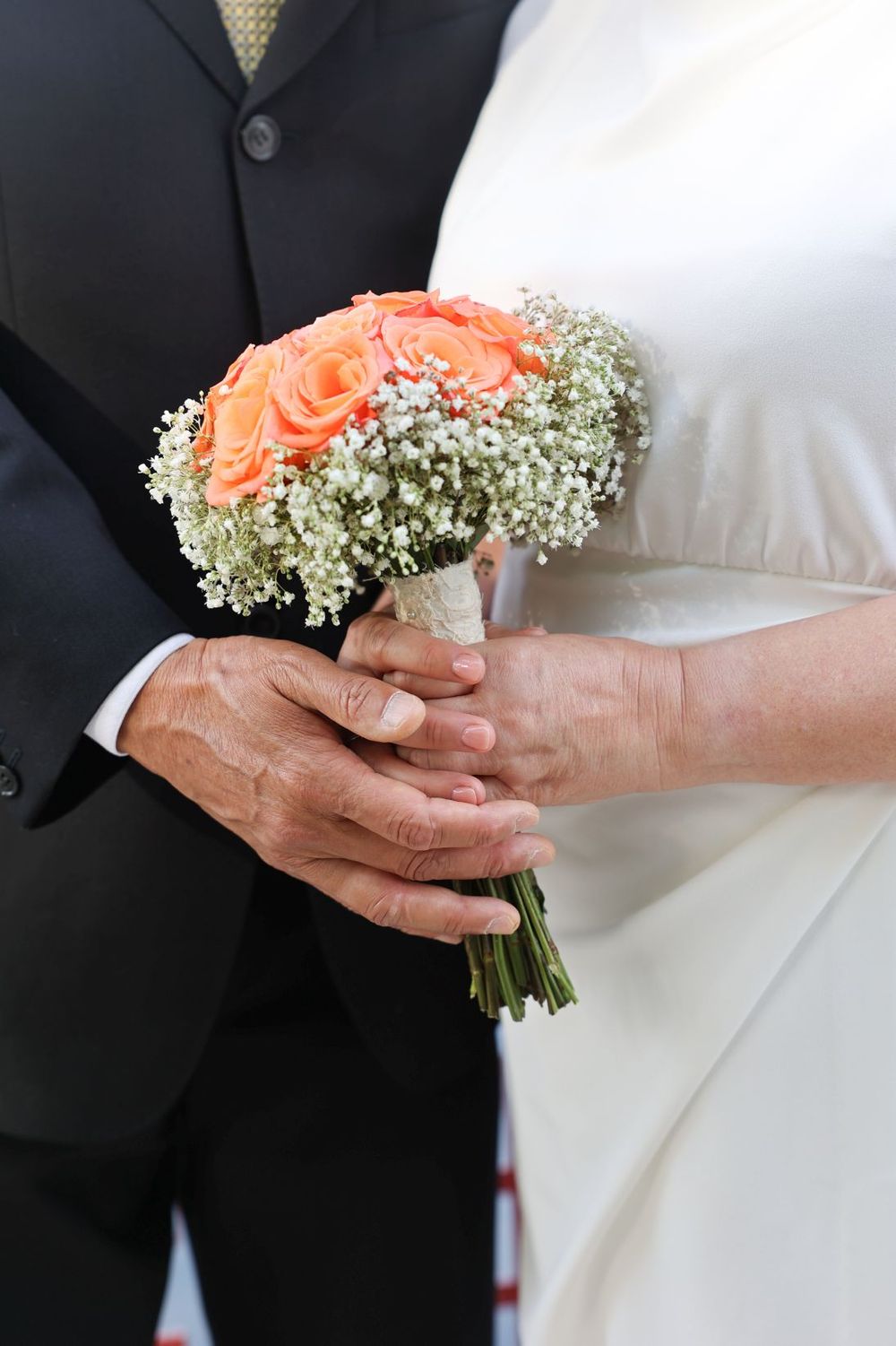 A wedding bouquet of peach roses and baby's breath held jointly by two hands in formal attire.
