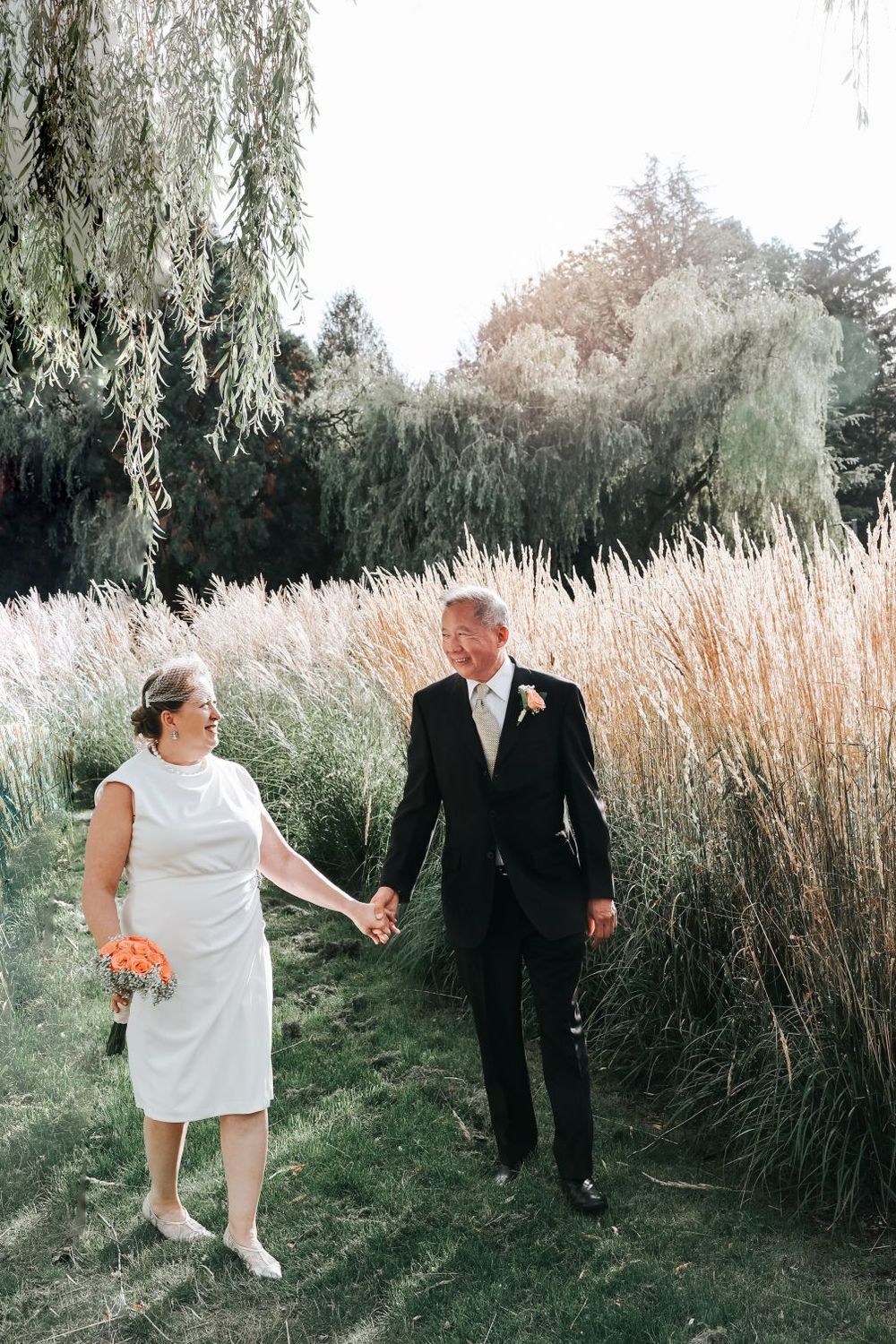 A couple walks hand in hand through tall ornamental grasses and willow trees in a garden setting.