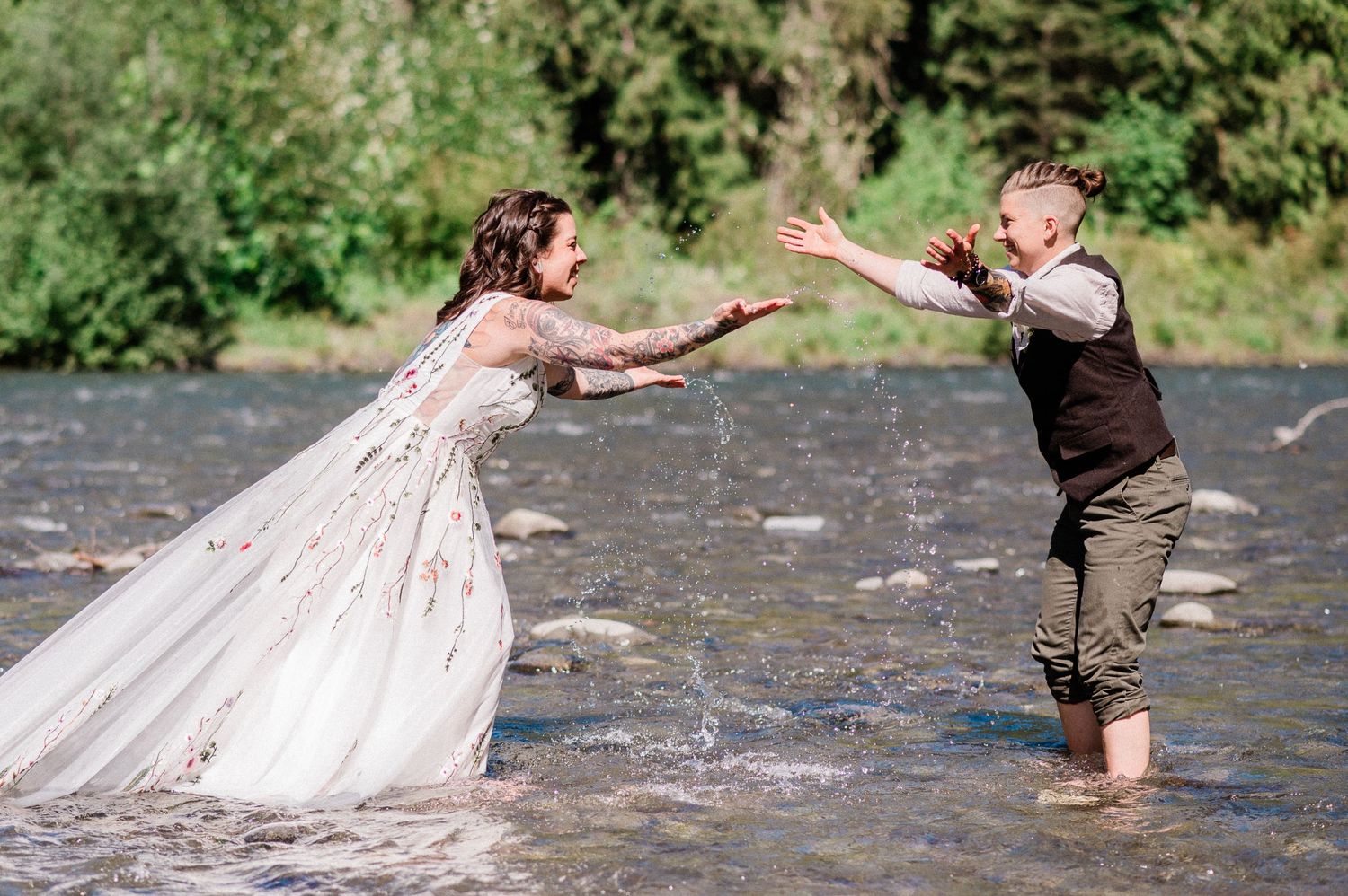 couple splashing water at each other while standing in river in their wedding attire