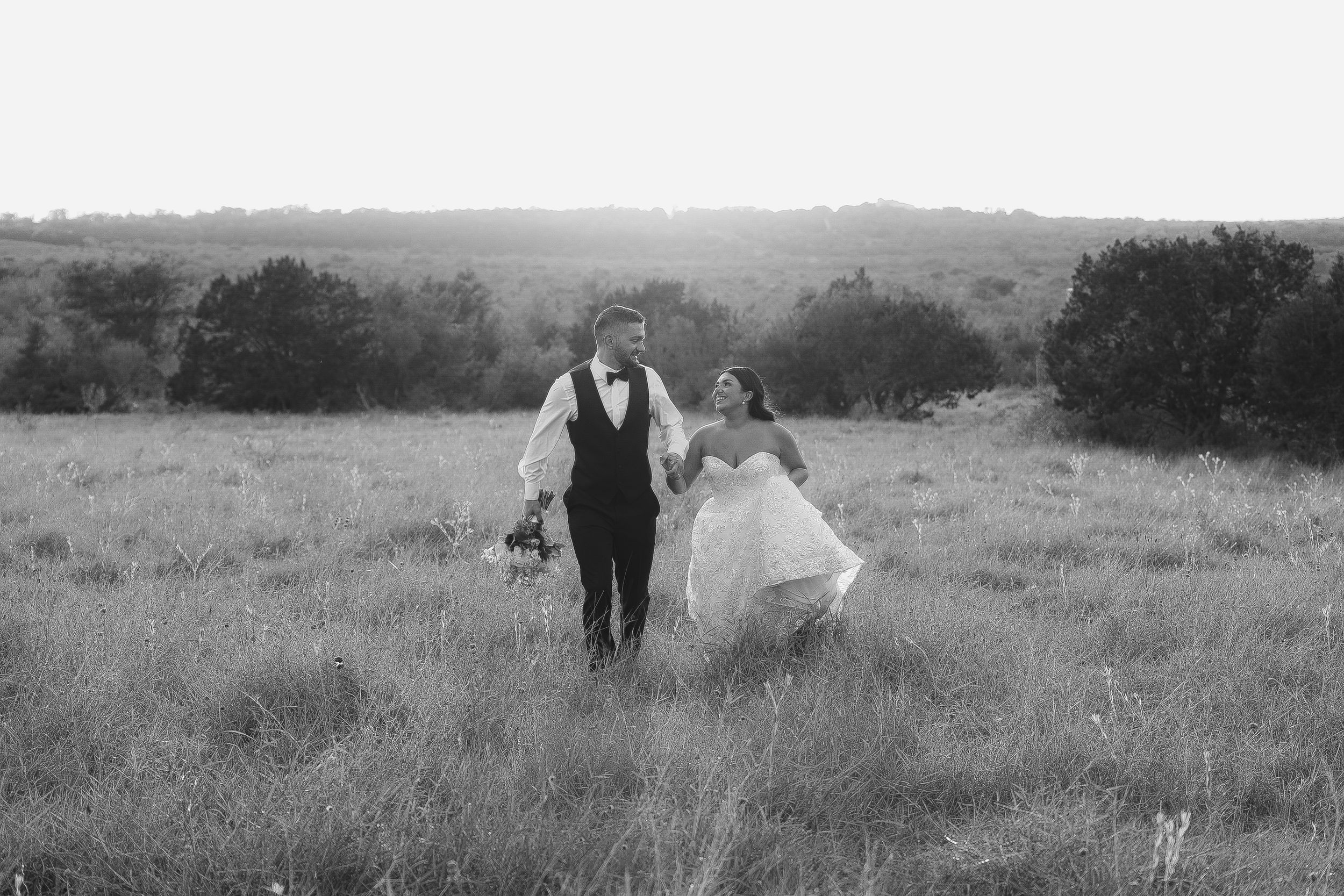 Black and white photo of couple walking through meadow holding wedding bouquet.