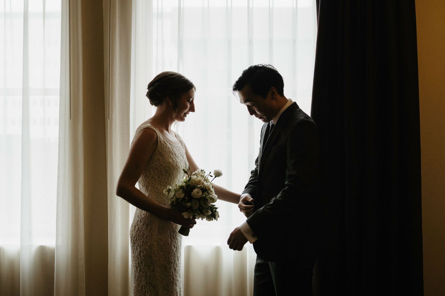 Bride and groom silhouetted by window light, bride holding white bouquet, groom adjusting cufflinks before wedding ceremony.