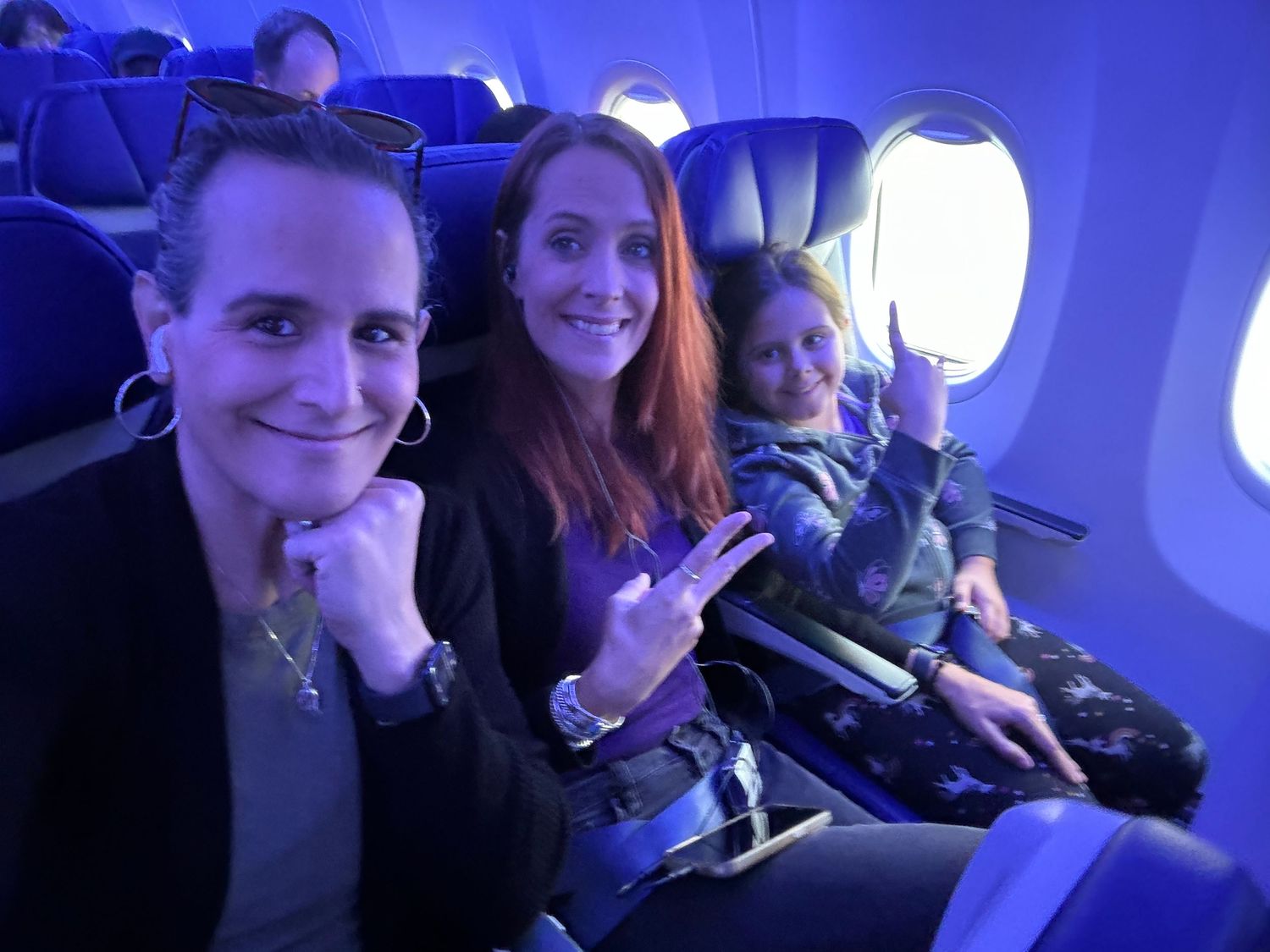 Three women smiling and posing for a selfie inside a JetBlue airplane with blue ambient lighting.