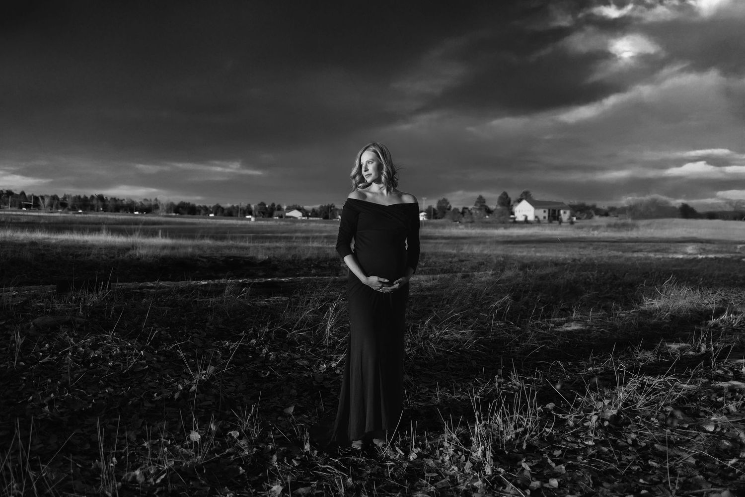 Black and white photo of a pregnant woman in a dark dress standing in an open field under dramatic cloudy skies.