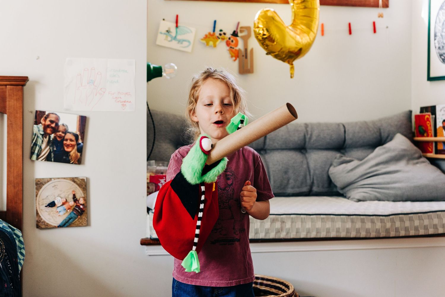 Young child playing with a toy dinosaur puppet on a cardboard tube in a cozy decorated bedroom.