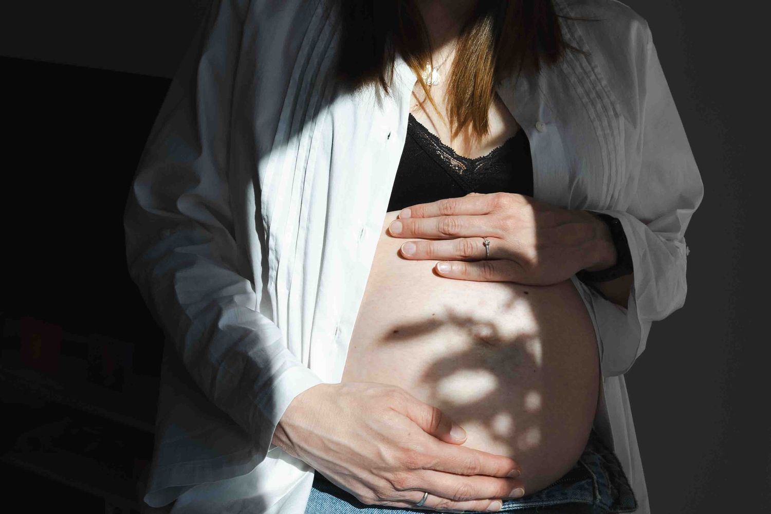 Pregnant woman in white shirt cradling her bare baby bump with both hands in soft natural light.