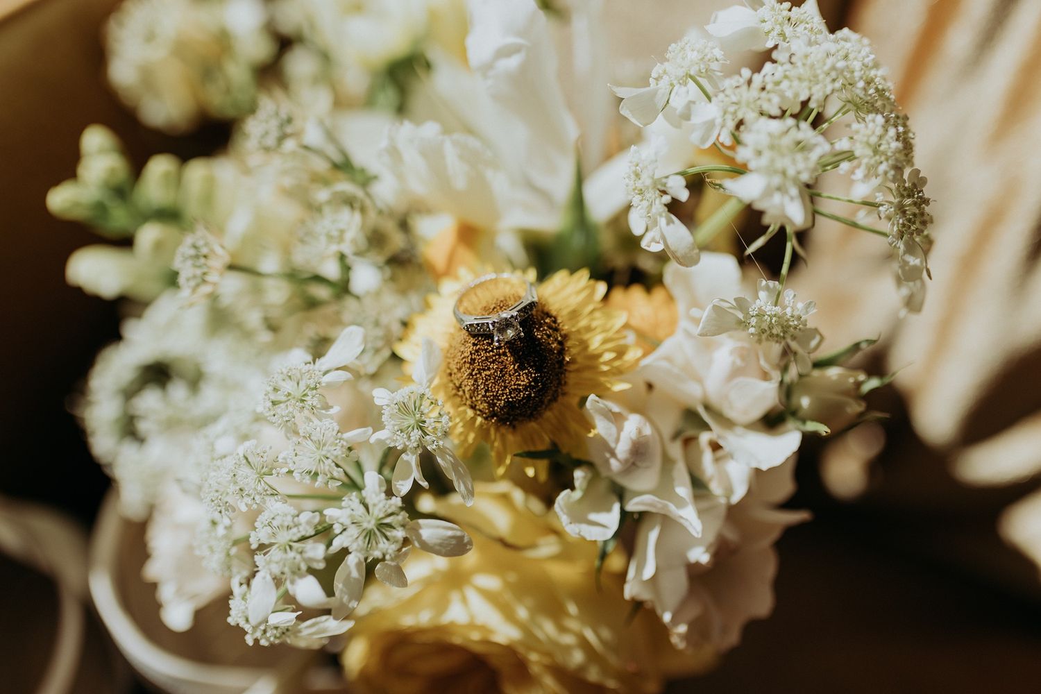 Close-up of a wedding bouquet with white blooms, yellow flowers, and two rings resting on a sunflower center at The Lane.