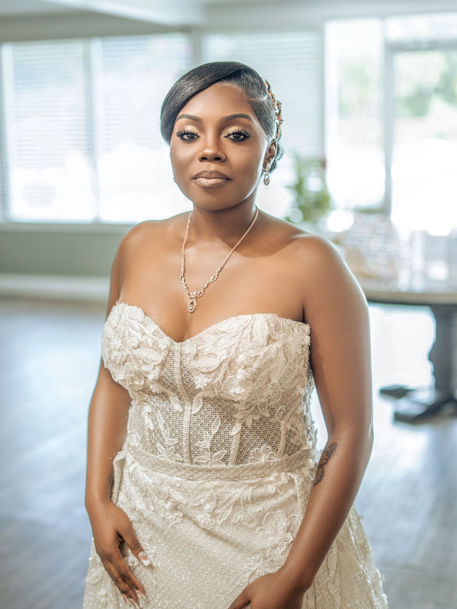 Elegant bride in a strapless lace wedding gown with hair accessories and necklace, posing indoors.