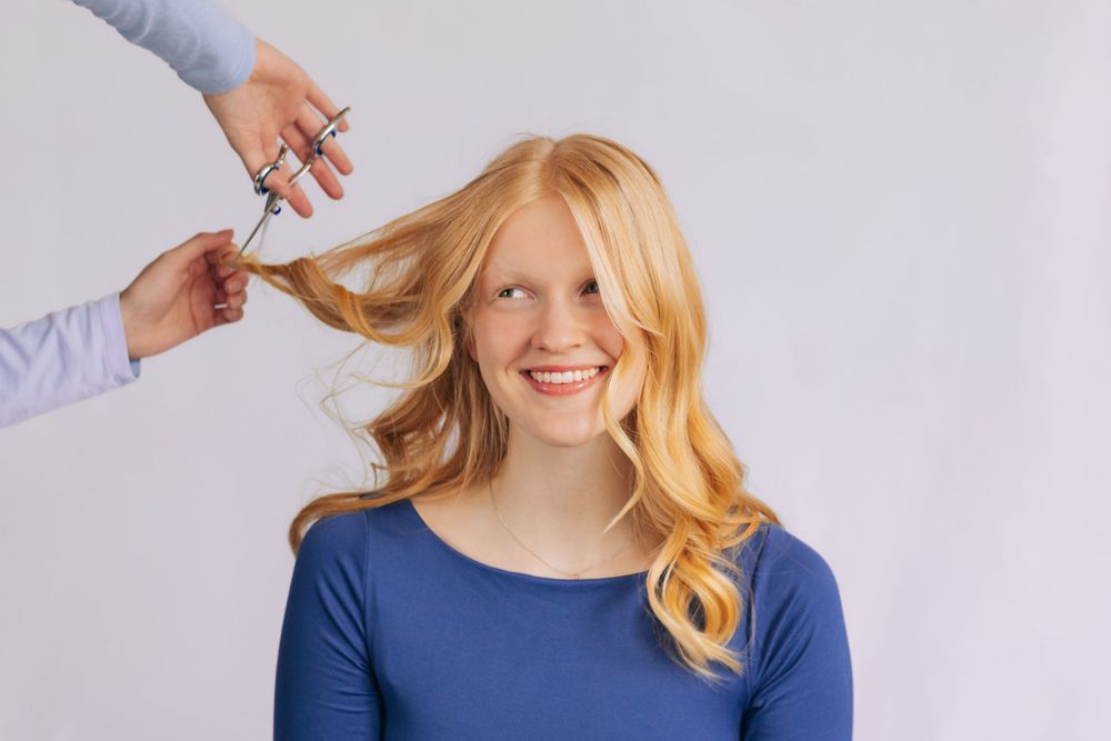 Smiling young woman with long blonde wavy hair getting a haircut at a salon against a white background.