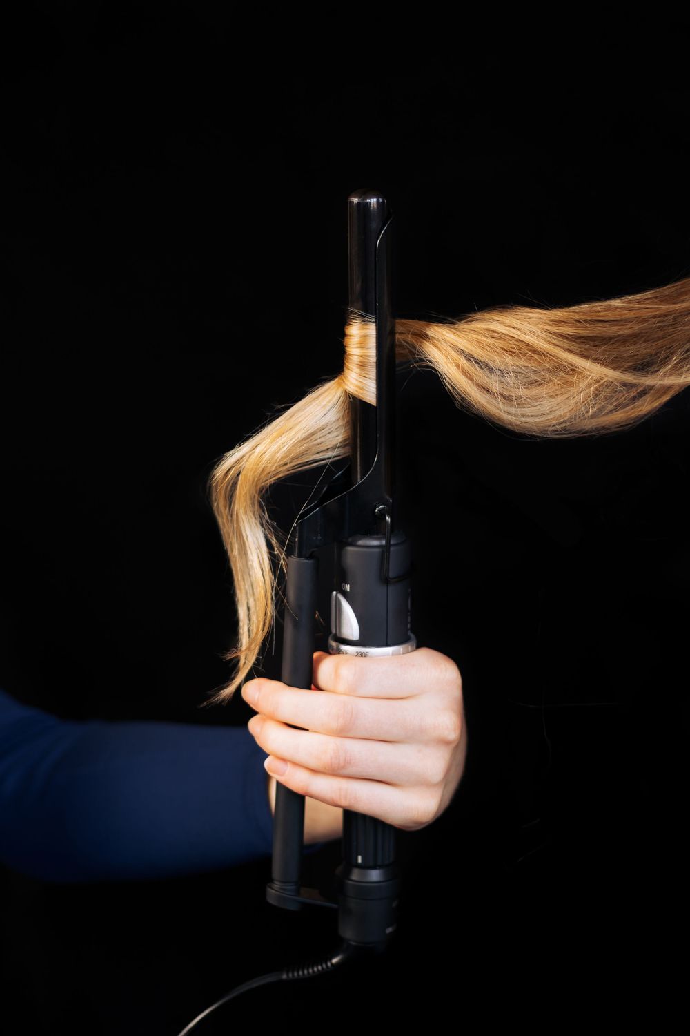 Hand holding a curling iron with blonde hair wrapped around the barrel against black background.
