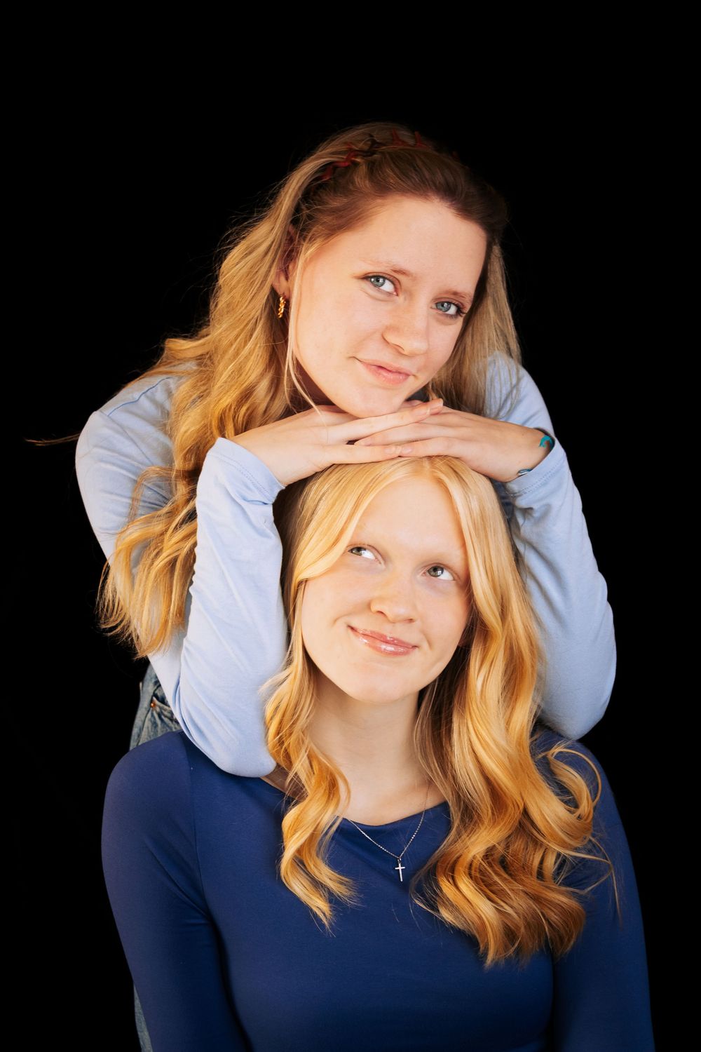 Two young blonde women posing together against black background, one resting hands on the other's head.