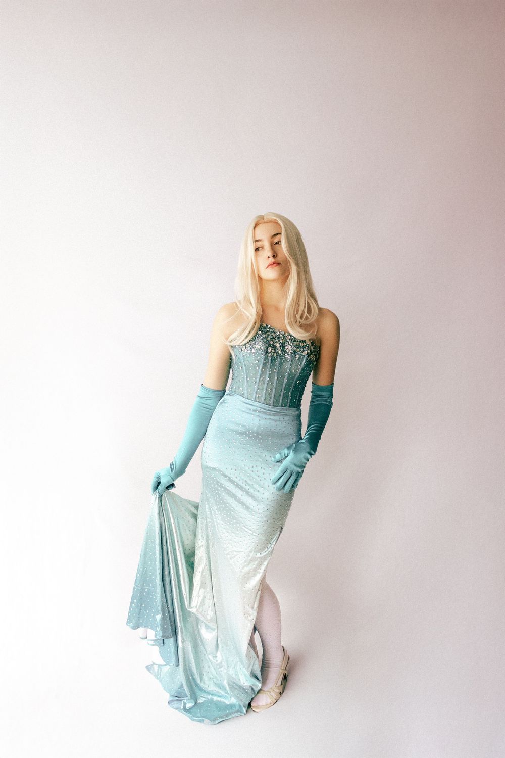 Woman in elegant teal beaded strapless gown with matching gloves poses gracefully against white background.