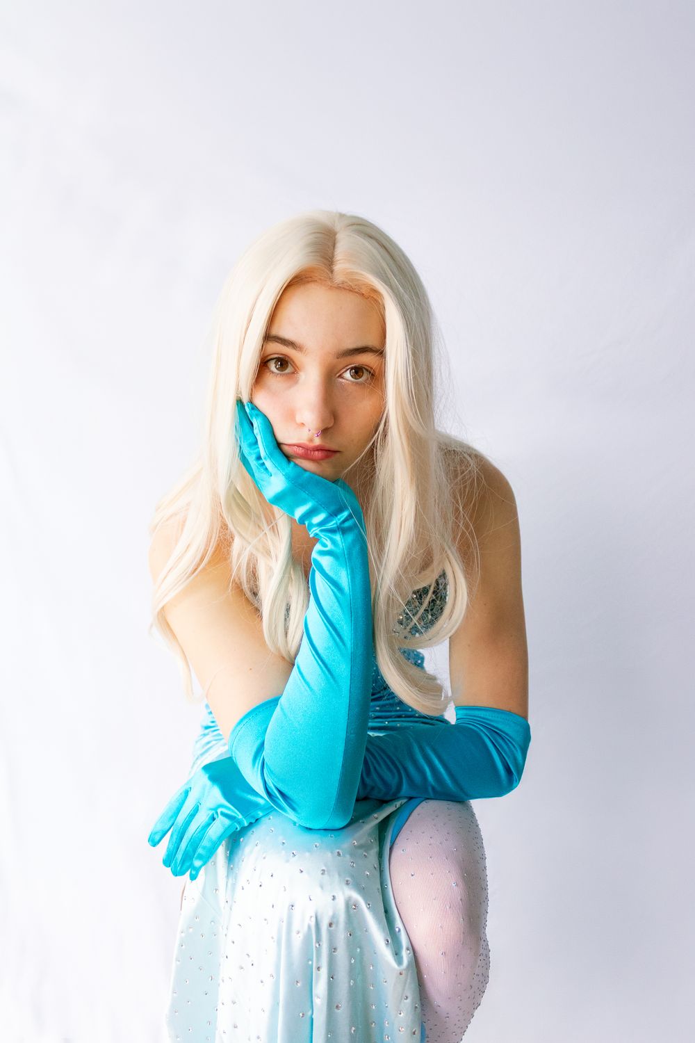 Young woman in blonde wig, blue satin gloves, and sparkly light blue dress poses against white background.