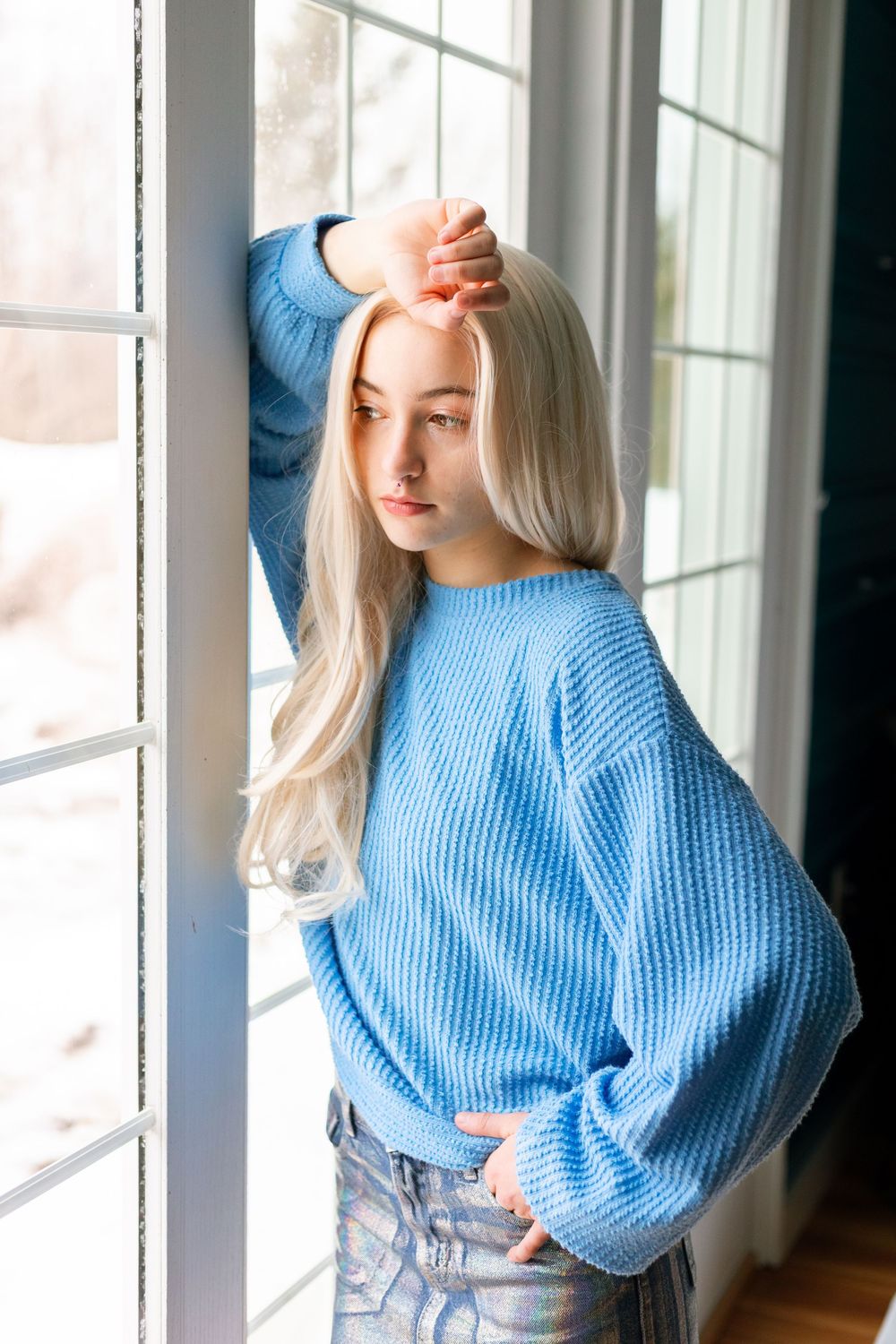 Young blonde woman in a blue knit sweater and jeans posing by a white window frame indoors.