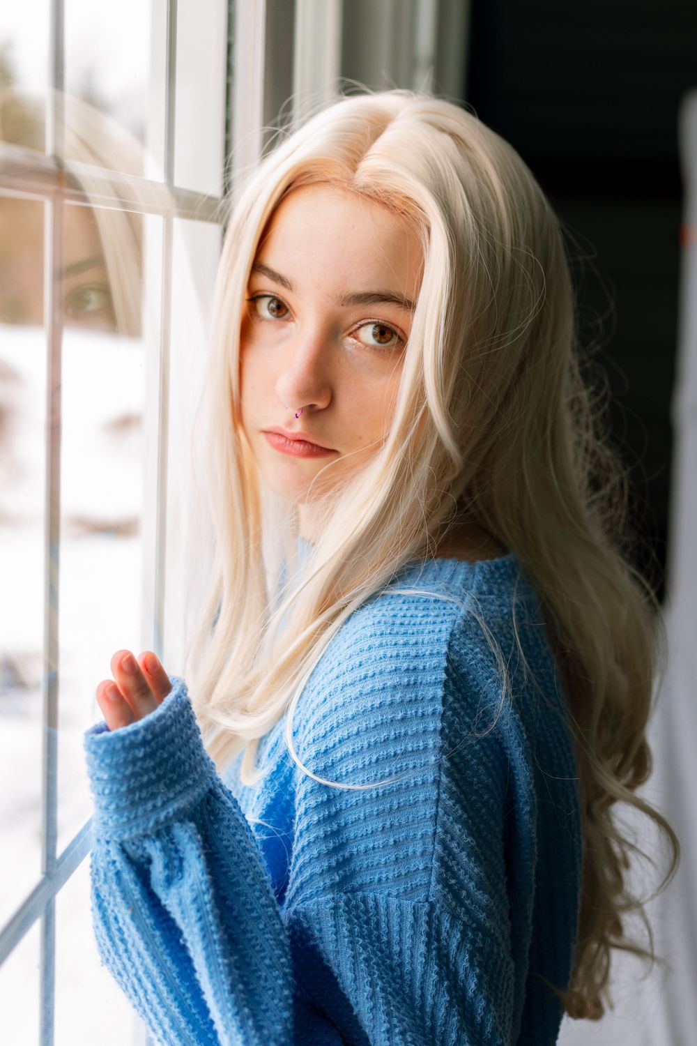 Young woman with long blonde hair wearing a blue knit sweater, posing by a window with snowy background.