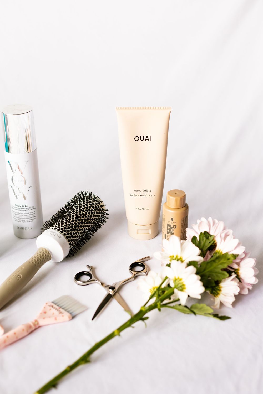 OUAI curl creme, round brush, scissors, and floral accents arranged on white fabric for a hair care flatlay.