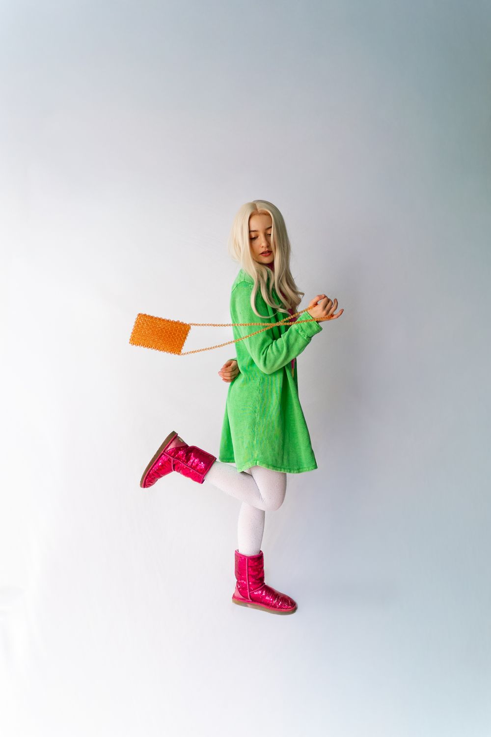 Woman in green mini dress, white tights, pink glitter boots, holding orange crossbody bag on white background.