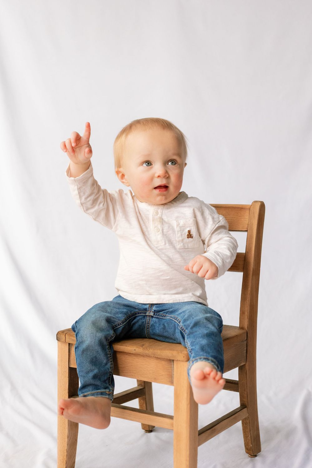 A cute baby in jeans and white shirt sits on a wooden chair, pointing upward with one hand raised.