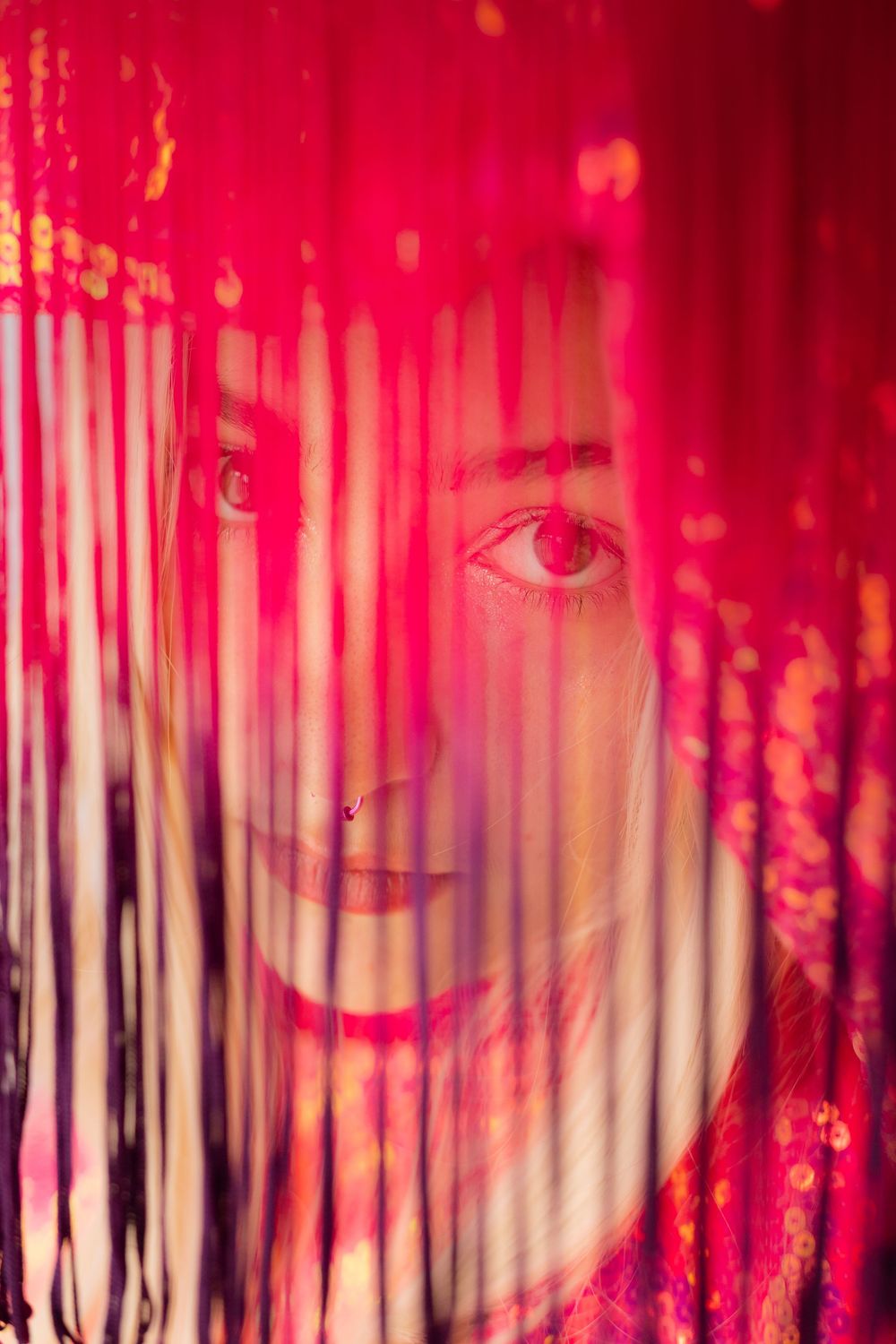 A smiling woman peers through vibrant red curtain strings, her face partially visible with a warm, mysterious glow.
