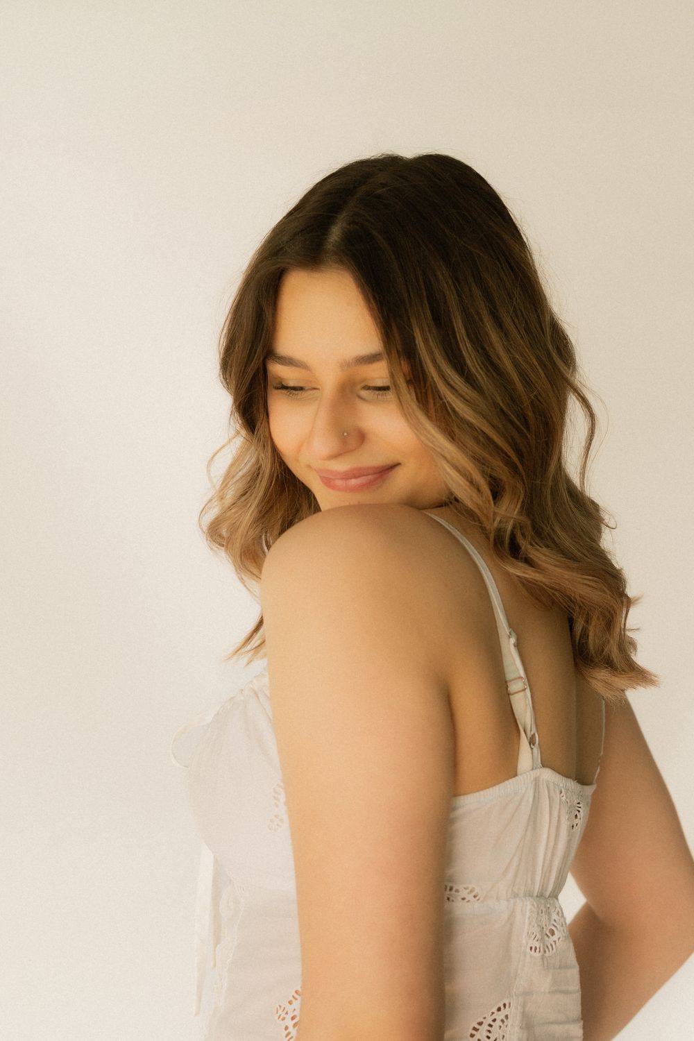 Young woman with wavy brown hair smiling softly, wearing a white sundress against a light background.