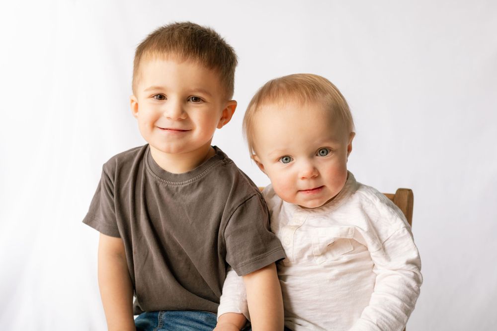 Two young siblings, a smiling toddler boy and a baby, pose together for a portrait against a white background.