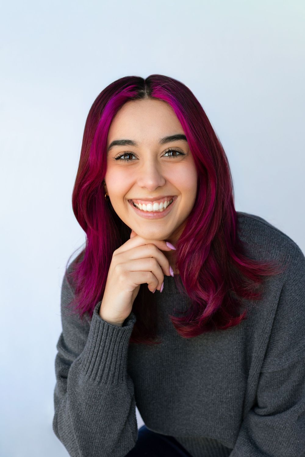 Young woman with vibrant pink-dyed hair smiling brightly, wearing a gray sweater, resting chin on hand against white background.