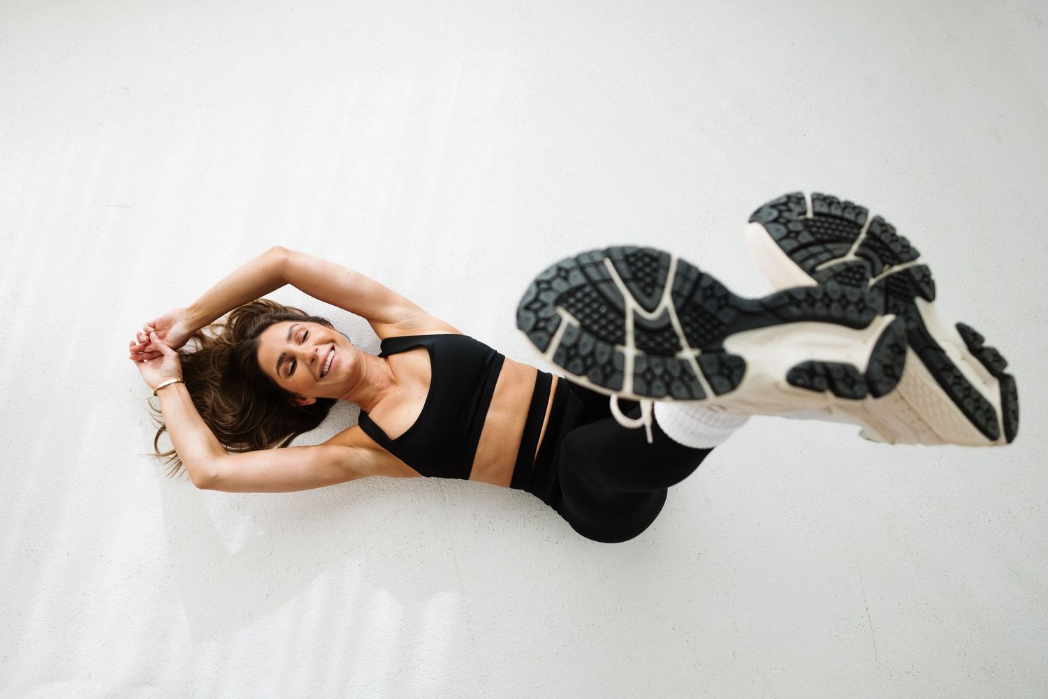 Woman in black sports bra lying on white floor stretching in patterned sneakers and leggings.