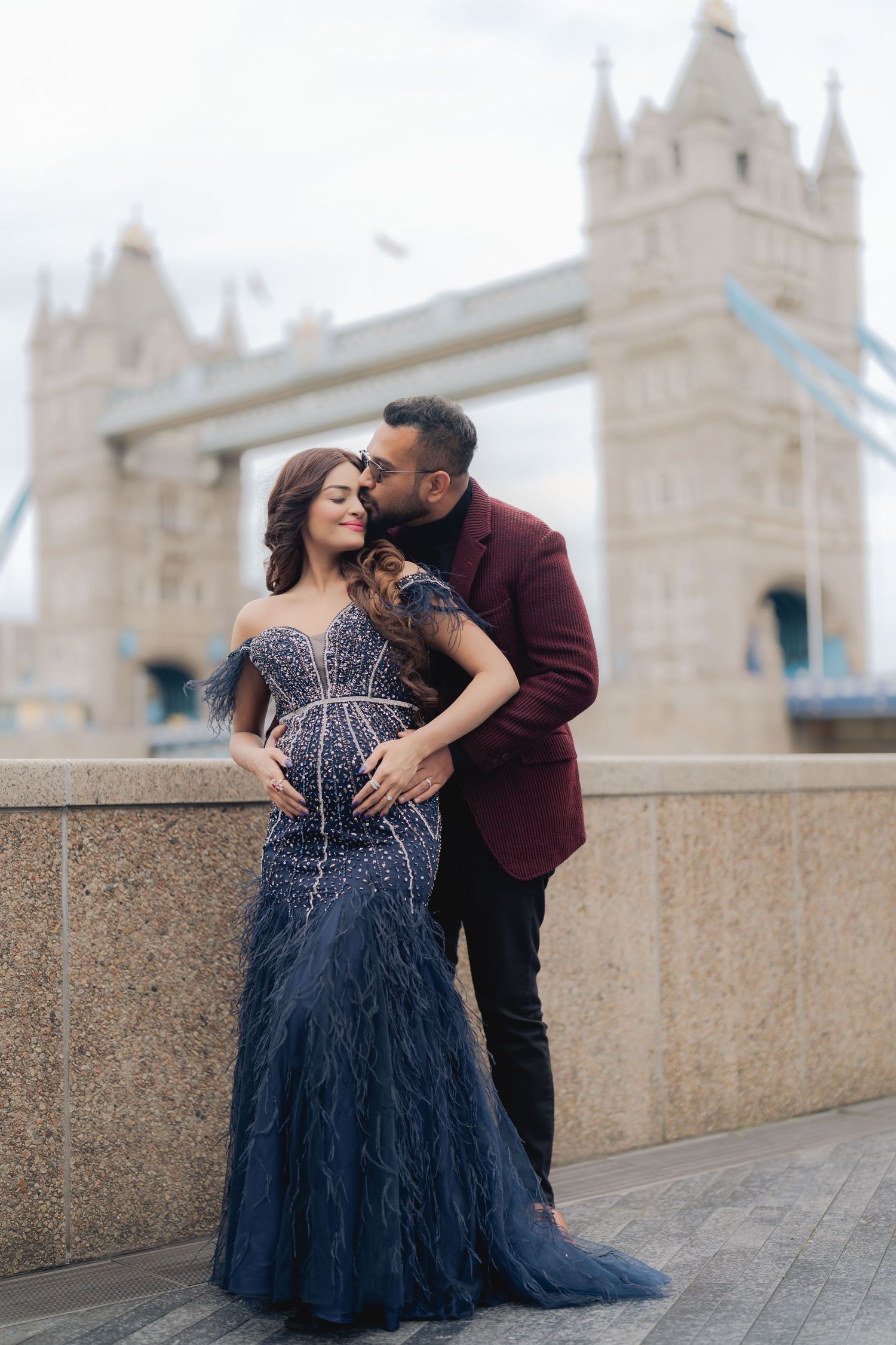 Couple sharing tender kiss before Tower Bridge London, pregnant woman in navy beaded gown, man in burgundy jacket.