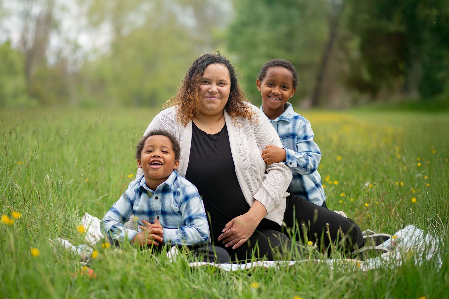 A mother sits in a green meadow with her two young sons, all smiling, wearing blue plaid shirts outdoors.