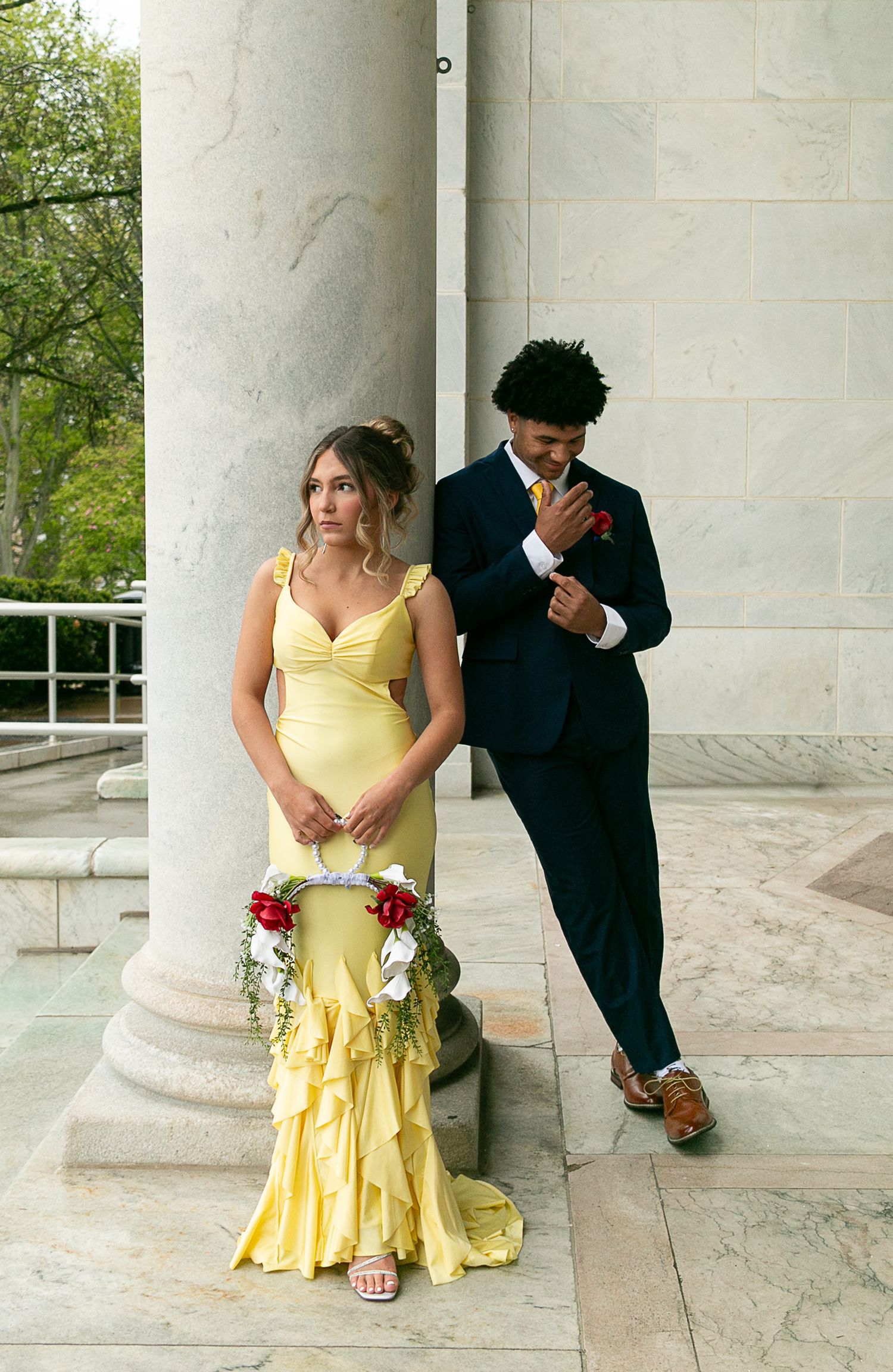 A young couple in prom attire, she in yellow gown with floral bouquet, he in navy suit, posing by marble columns.