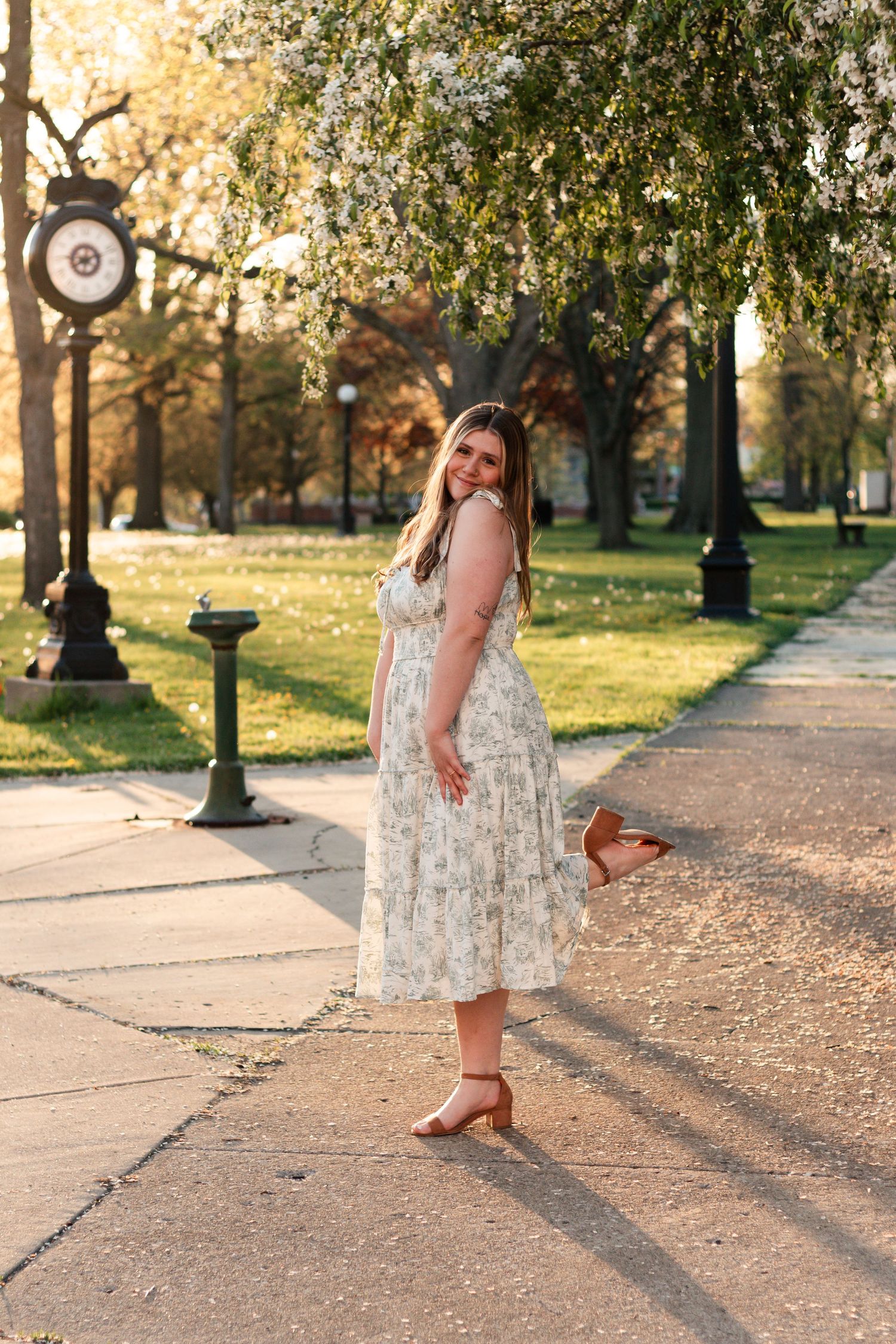 Woman in floral midi dress and heels posing in a sunny park with blooming white trees in spring.