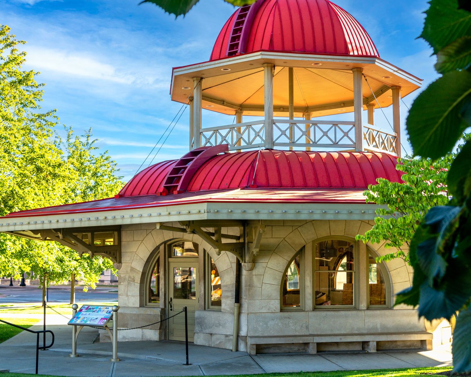 Historic stone gazebo with red dome roof in a sunny park setting with lush green trees and blue sky.