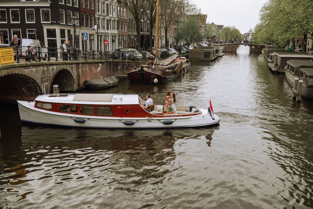 A white and red boat cruises along an Amsterdam canal with historic buildings and moored houseboats in the background.