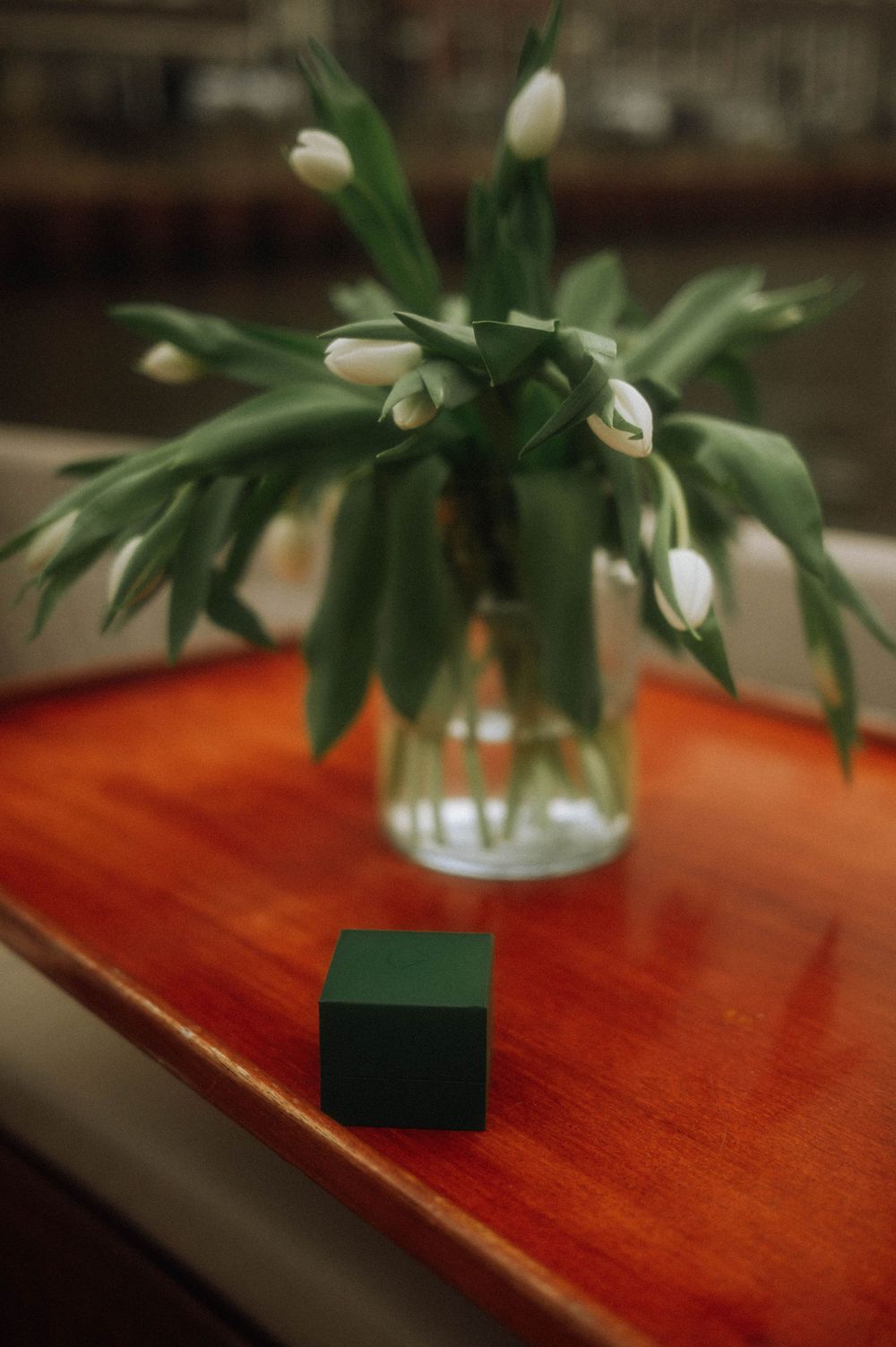 A small green ring box sits on a wooden table beside a glass vase of white tulips. Secret proposal Amsterdam photoshoot