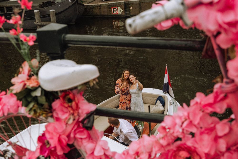 Two women posing on a boat in Amsterdam canal surrounded by pink flowers with a Dutch flag visible.