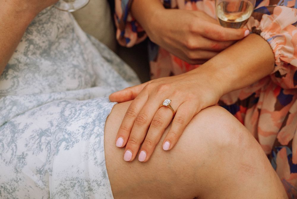 Close-up of a woman's hand with pink nails wearing a diamond engagement ring resting on her knee.
